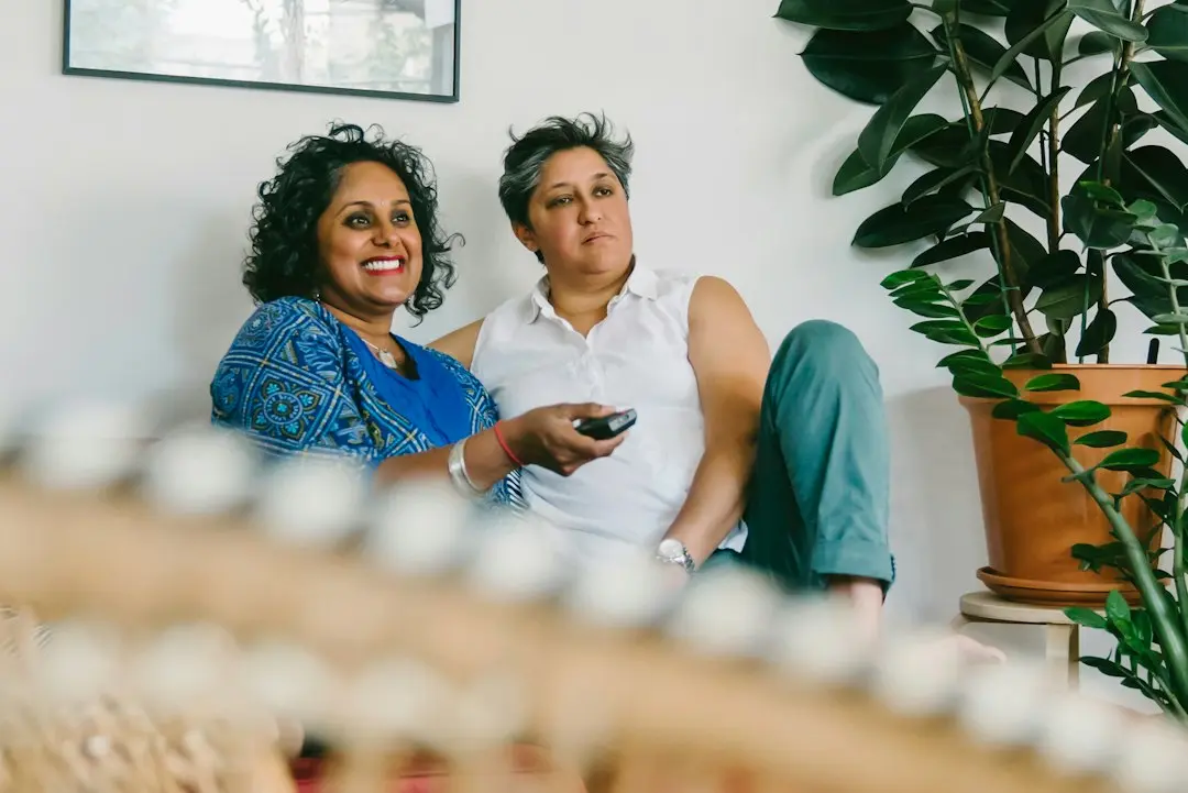 Two women relaxing on a sofa, one pointing a remote at the TV, as when browsing the Glomax8K TV guide